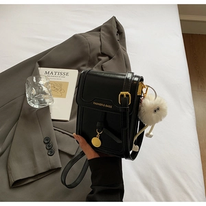 Close-up of black glossy leather crossbody bag with gold buckle and plush pom-pom charm