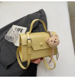 Detail of yellow rectangular crossbody bag with teddy bear charm and striped ribbon accent