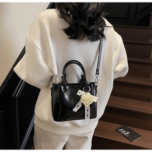 Close-up of black glossy tote bag featuring a pearl lace bow charm and crossbody strap