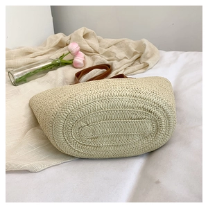 Detailed close-up of woven straw tote bag with brown leather handles on white fabric