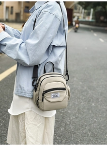 Detail of compact beige canvas shoulder bag featuring black zippers and woven logo label