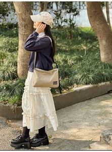 Outdoor shot of a beige canvas crossbody bag with black wide strap and logo detail