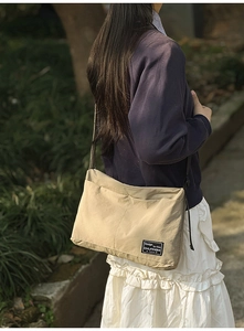 Side view of a khaki nylon crossbody bag with brand label in a natural garden setting