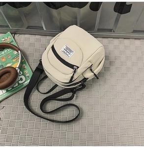 Flat lay view of a compact cream nylon crossbody bag with black adjustable strap and zippers
