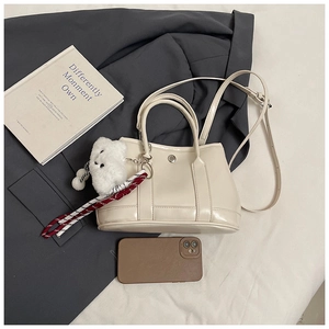 Detail view of cream PU leather handbag with metal buckle, plush charm, and phone for scale