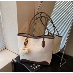 Detail of cream tote bag with brown trim, gold logo, and fluffy pendant in home setting