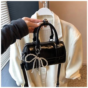 Close-up of black patent leather pillow bag featuring a pearl bow pendant and shoulder strap