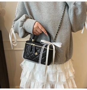 Detail of black quilted leather box bag with gold floral logo and white lace bow