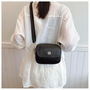 Close-up of a black crossbody bag with round metal logo worn by a model against white wall