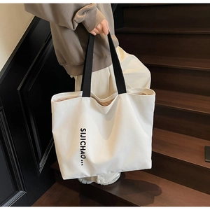 Close-up of a large beige tote bag with black shoulder straps and vertical letter print