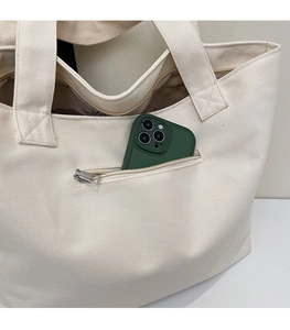 Detail of white canvas tote bag showing the front zipper pocket holding a large smartphone