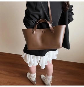 Brown leather tote bag worn as a shoulder bag with a black suit and white pleated skirt