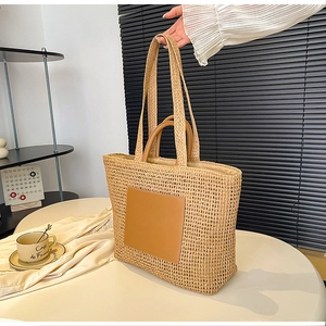 Detail of a beige woven straw tote bag featuring a brown faux leather square patch and handles