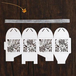 Flat lay of laser-cut white wedding favor box with tropical leaf patterns and silver ribbon