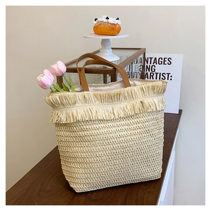 Detail of straw woven tote bag with fringe trim and brown leather handle on a wooden table
