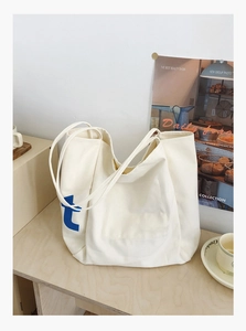 Détail du sac cabas en toile crème montrant la lettre 't' bleue et la poche sur la table
