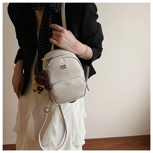 Détail du sac à dos blanc cassé avec texture litchi, logo métal et breloque chien