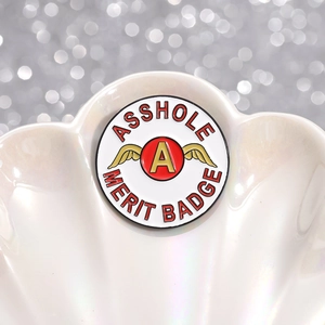 Lustiger 'Asshole Merit Badge' Emaille-Pin mit Goldflügeln in einer weißen Muschel