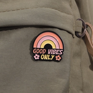 Détail d'un pin's émaillé arc-en-ciel avec texte 'Good Vibes Only' sur toile olive