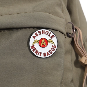 Makro-Detail des Emaille-Pins 'ASSHOLE MERIT BADGE' mit Goldflügeln auf Leinentasche