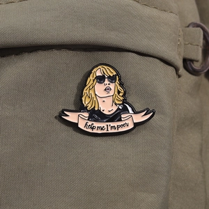 Close-up of enamel pin with sunglasses character and Help Me I'm Poor pink banner