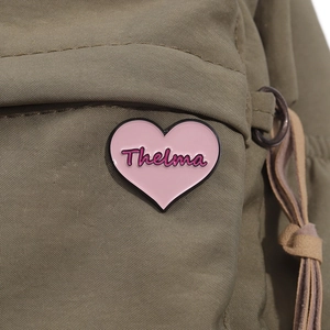 Close-up of pink heart enamel pin with Thelma engraving on olive green canvas bag