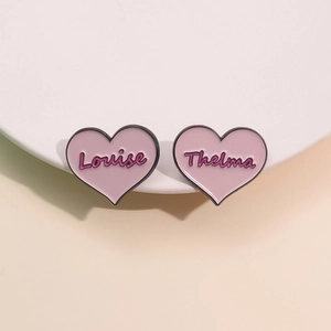 Pink heart-shaped enamel pins set with Louise and Thelma script lettering on white tray