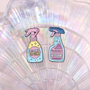 Enamel pins shaped like spray bottles with BE-GONE and REPELLENT slogans on iridescent tray