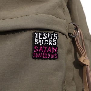 Close-up of black enamel pin with Jesus and Satan text on olive green canvas fabric