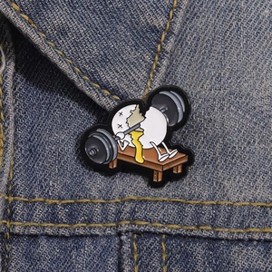Funny cartoon enamel pin showing an egg character struggling with a heavy barbell on denim