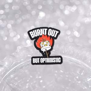 Macro shot of an enamel pin featuring a burnt marshmallow and optimistic quote slogan