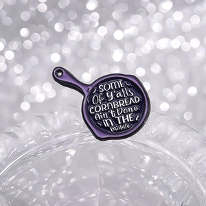 Gros plan d'un pin's en émail violet en forme de poêle avec citation sur verre ondulé