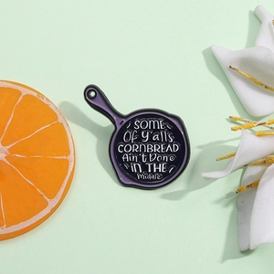 Purple skillet enamel pin with funny cornbread quote on a green background with props