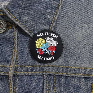 Cartoon enamel pin with flowers and Pick Flowers Not Fights slogan on denim jacket