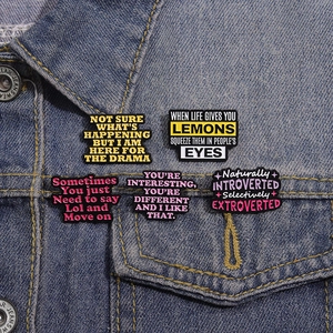 Five assorted colorful enamel pins with funny slogans displayed on a blue denim jacket