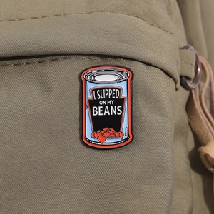 Blue enamel bean can brooch with 'I SLIPPED ON MY BEANS' text on khaki canvas bag