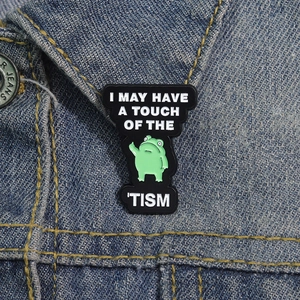 Dettaglio della spilla a rana con slogan I May Have A Touch Of The Tism su giacca denim