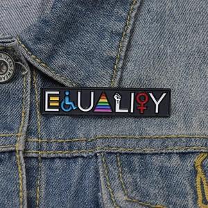 Detail of Equality enamel pin featuring rainbow and social symbols on denim jacket