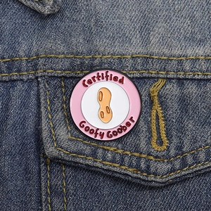 Round pink enamel pin with peanut design and Certified Goofy Goober text on denim