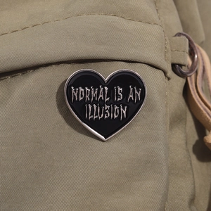 Enamel heart pin 'NORMAL IS AN ILLUSION' on a canvas backpack strap