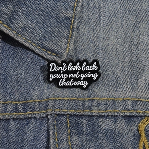 Black enamel brooch with 'Don't look back' quote pinned on a blue denim jacket