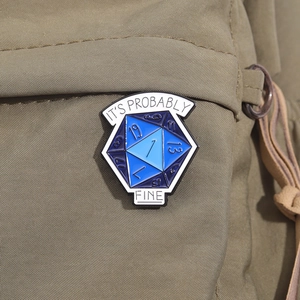 Macro detail of blue D20 enamel pin with It's Probably Fine text on green canvas