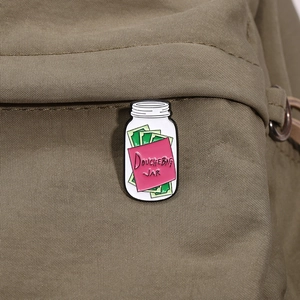 Détail d'un pin's émaillé en forme de pot avec texte Douchebag Jar sur sac à dos olive