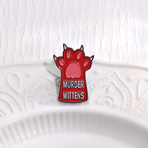 Red cat paw enamel pin with Murder Mittens text on a white textured background