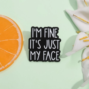 Black enamel brooch featuring white text I'M FINE IT'S JUST MY FACE on a green background