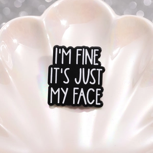 Black enamel brooch with 'I'M FINE IT'S JUST MY FACE' quote on a white ceramic tray