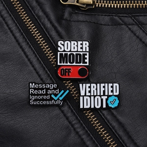 Detail von lustigen Emaille-Pins mit SOBER MODE OFF-Text auf einer schwarzen Lederjacke