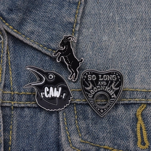 Three gothic enamel pins featuring a black goat, a crow, and a planchette on denim fabric