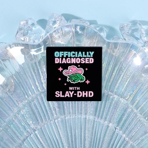 Détail d'un pin's carré en émail avec un motif de grenouille et texte Slay-DHD stylé