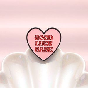 Rosa Emaille-Anstecker in Herzform mit Good Luck Babe Schriftzug auf Muschelhintergrund